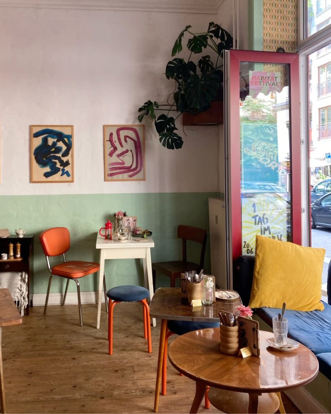 Farbenfrohe Café-Ecke mit mintgrüner Wand, orangefarbenen Stühlen, abstrakter Kunst, Holztischen und Tageslicht am Fenster